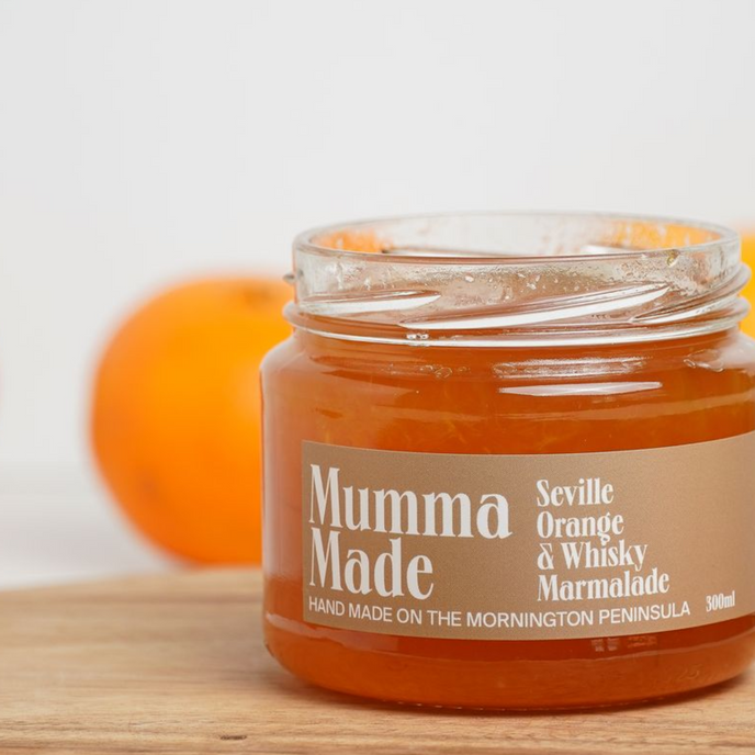 Jar of Mumma Made Seville Orange & Whisky Marmalade on a wooden surface with an orange in the background.