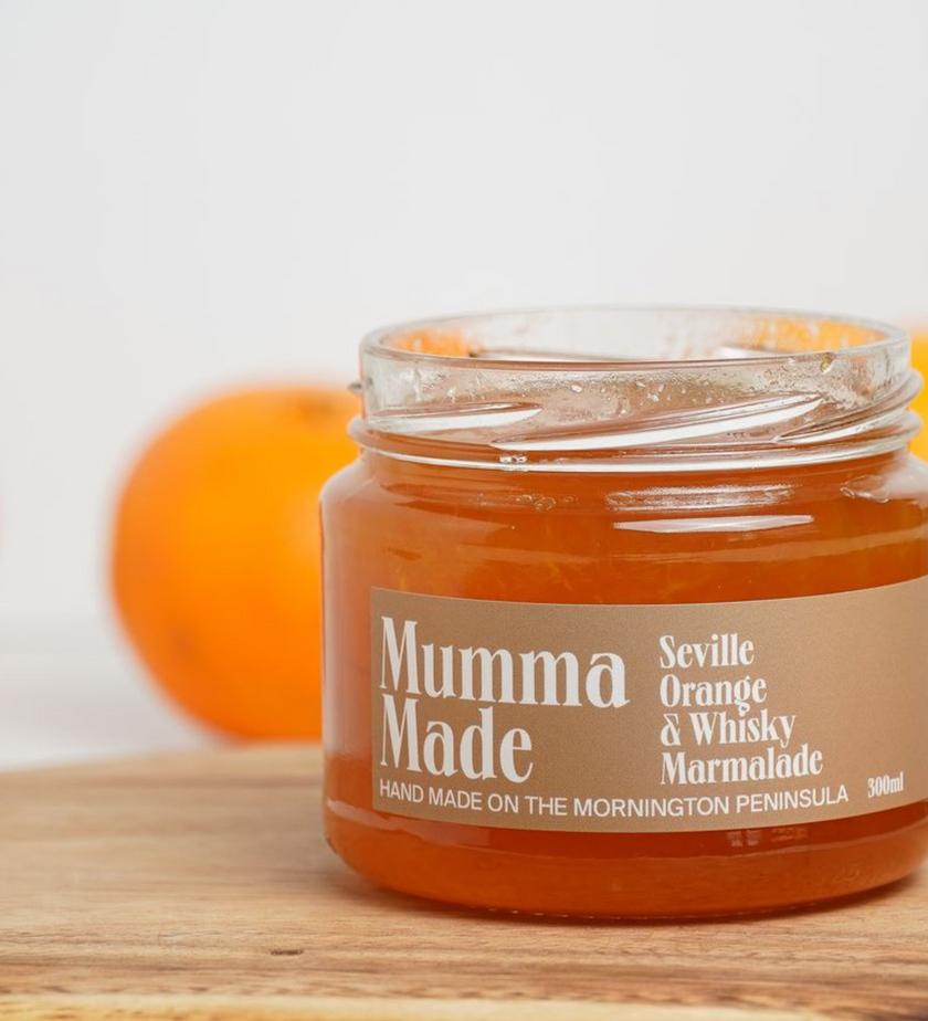 Jar of Mumma Made Seville Orange & Whisky Marmalade on a wooden surface with an orange in the background.