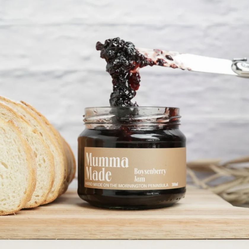 A jar of boysenberry jam with a slice of bread being dipped into it, placed on a wooden board with a knife and additional bread slices in the background.