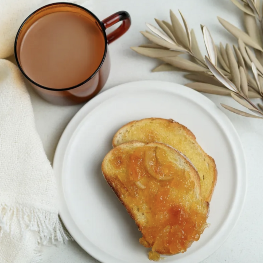 A jar of breakfast marmalade on a white plate with a piece of toast, alongside a cup of tea and a decorative branch in the background.