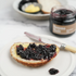 A slice of bread with blackberry jam on a plate, with a jar of blackberry jam and a knife in the background.