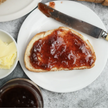 A piece of toast with strawberry and rhubarb jam on top, served on a white plate with a knife and a small bowl of butter.