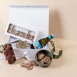 Open gift box with cookies, sprinkles, and a bottle of wine on a beige surface.
