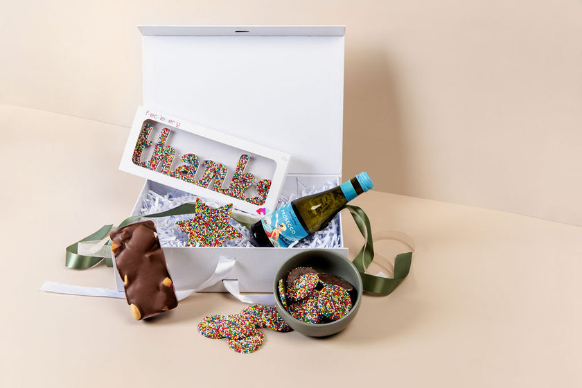 Open gift box with cookies, sprinkles, and a bottle of wine on a beige surface.