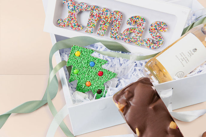 Decorative Christmas-themed gift box with a green tree and colorful letters on a beige background.