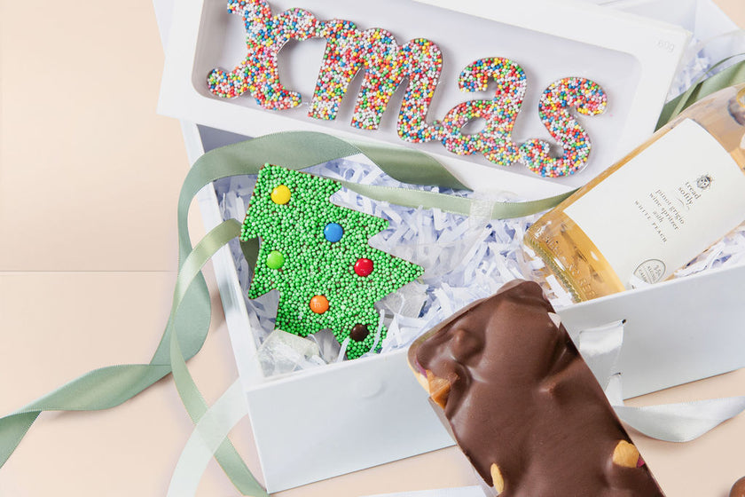 Decorative Christmas-themed gift box with a green tree and colorful letters on a beige background.