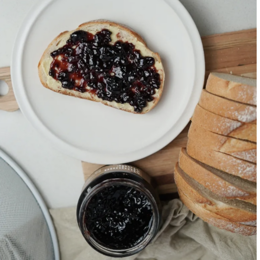 Toast with blackberry jam on a white plate, next to a jar of jam and a loaf of bread.
