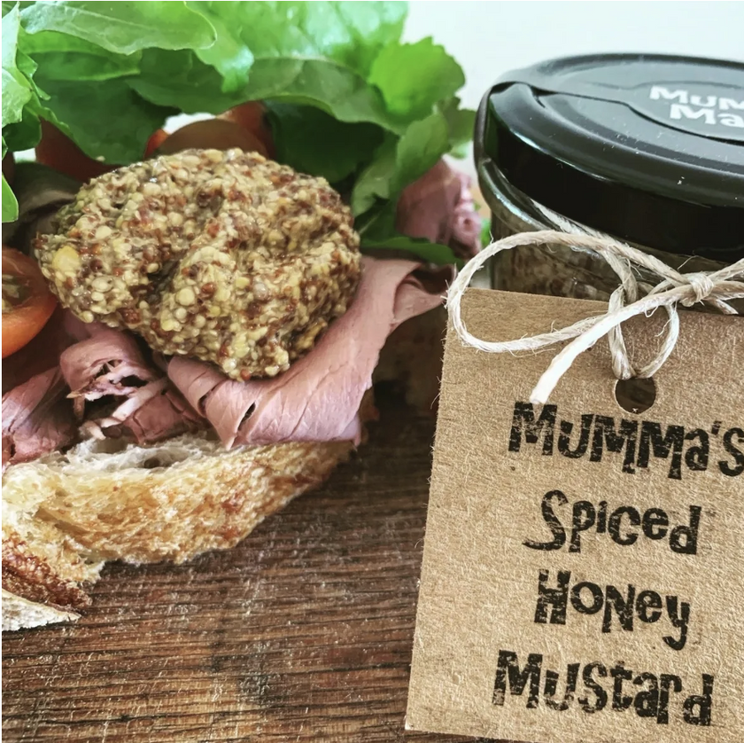 Sandwich with meat and bread, accompanied by a jar of 'Mumma's Spiced Honey Mustard' on a wooden surface.