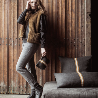 Woman in a brown vest and jeans standing against a wooden wall with a sofa and pillows in the foreground.