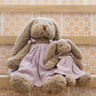 Two teddy bunny in pink purple dresses sitting on a wooden surface with a patterned background. Soft cuddly bunny for newborns or kids to cuddle and treasure. 