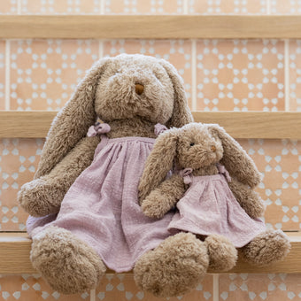 Two teddy bunny in pink purple dresses sitting on a wooden surface with a patterned background. Soft cuddly bunny for newborns or kids to cuddle and treasure. 