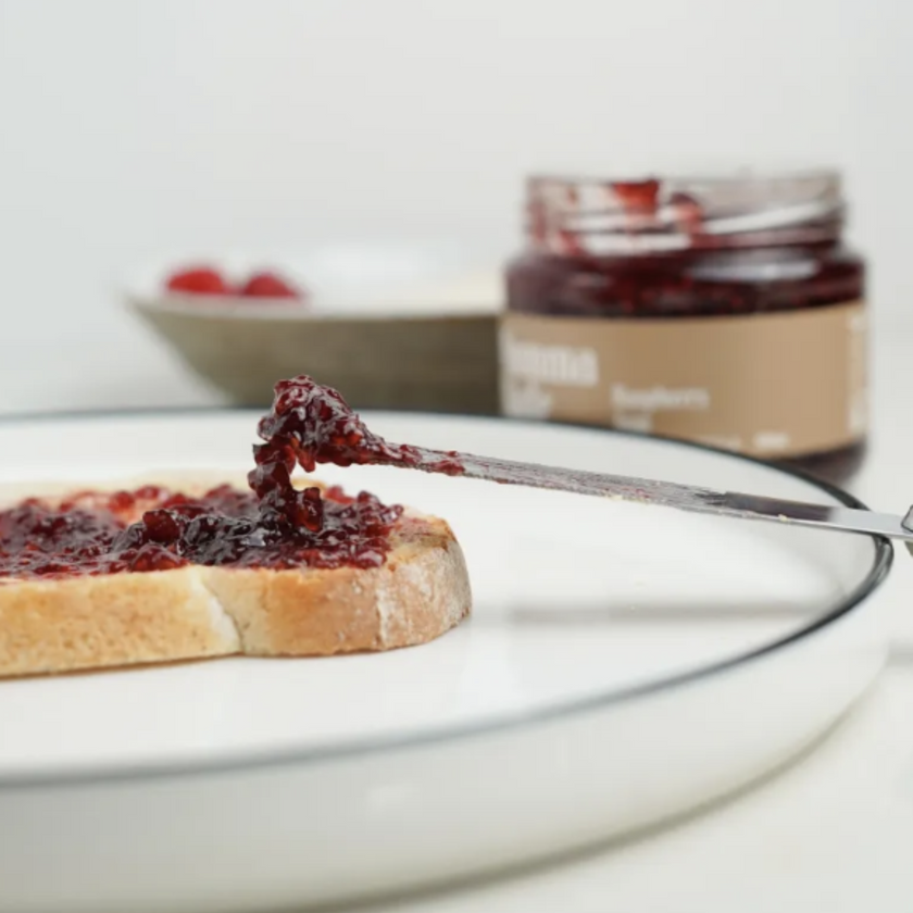 A slice of bread with raspberry jam on it, placed on a plate, with a jar of raspberry jam and a bowl of raspberries in the background.