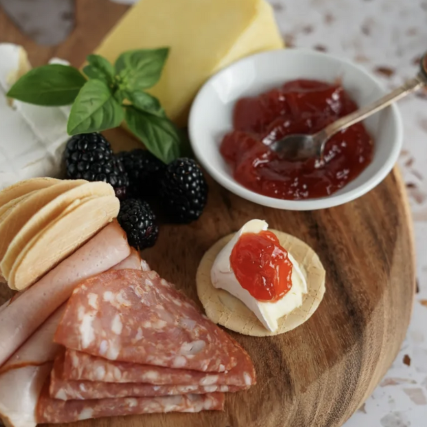 Wooden board with cured meats, blackberries, a wheel of cheese, and a small bowl of red jelly.