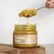 A spoon lifting green tomato pickle from a glass jar labeled 'Mumma Made'.
