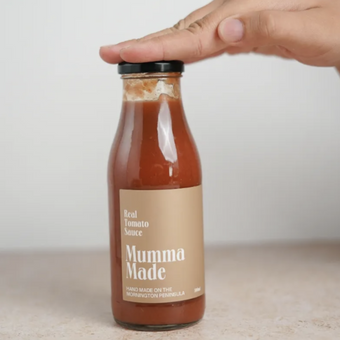 A hand holding a bottle of Real Tomato Sauce labeled 'Mumma Made' with a beige background.